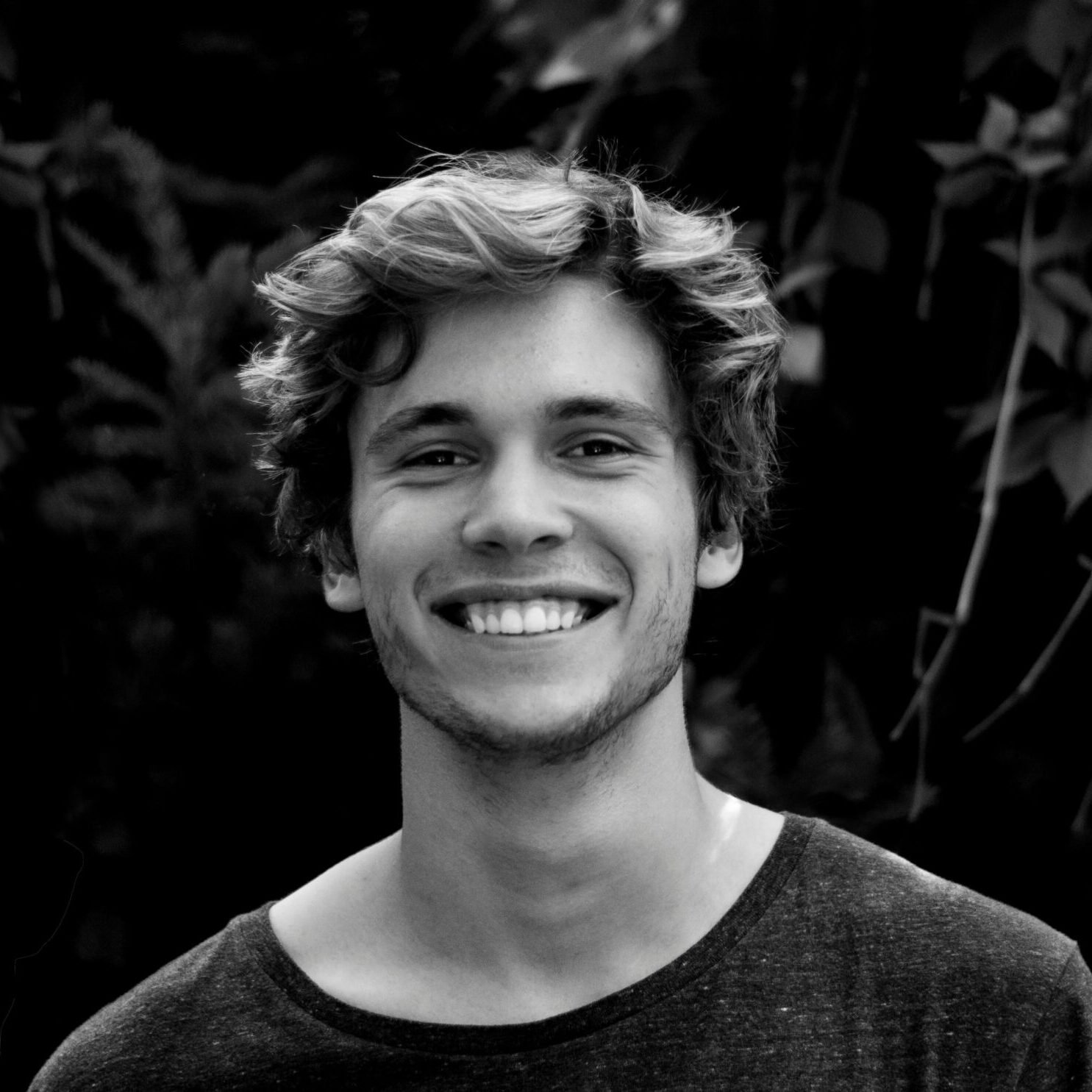 A black and white portrait of a young man smiling outdoors with natural background.